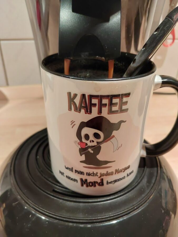 Kaffeetasse unter einer Senseo-Kaffeemaschine. Aufdruck auf der Tasse: Kaffee - weil man nicht jeden Morgen mit einem Mord beginnen kann.